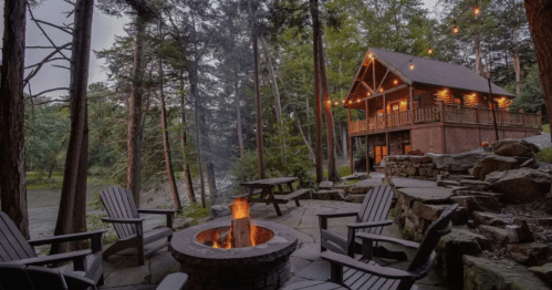 Cozy cabin in the woods with a fire pit, surrounded by trees and a serene river view. Warm lights illuminate the scene.