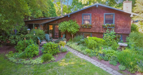 A charming wooden house surrounded by lush greenery and colorful flowers, with a stone pathway leading to the entrance.