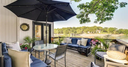 A cozy outdoor patio with a round table, chairs, and a large umbrella, overlooking a serene lake and greenery.