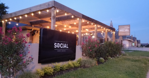 Exterior of a restaurant named "SOCIAL" with string lights, flowers, and a cozy atmosphere at dusk.