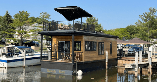 A floating houseboat with a rooftop deck, docked by the shore, surrounded by trees and boats on a calm waterway.