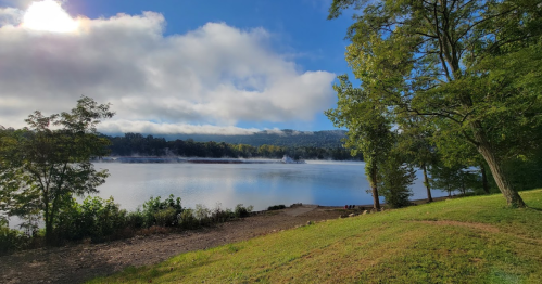 A serene lake surrounded by trees, with mist rising from the water under a blue sky and fluffy clouds.