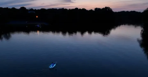 A serene river at dusk with a lone boat gliding across the calm water, surrounded by trees and a colorful sky.