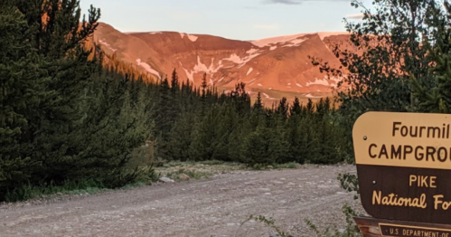 Sign for Fourmile Campground with a scenic mountain backdrop and trees under a sunset sky.
