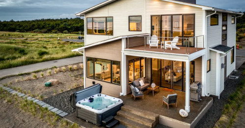 A modern house with large windows, a deck, and a hot tub, surrounded by greenery and a pathway.