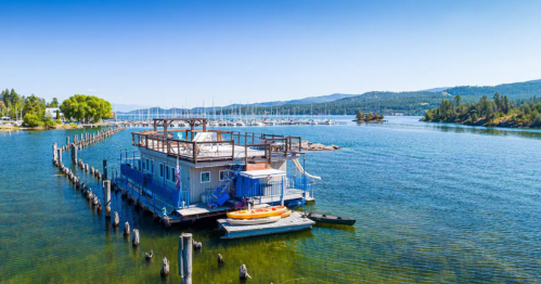 A blue houseboat docked by a marina, surrounded by calm water and green hills under a clear blue sky.