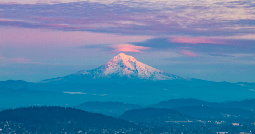 Snow-capped mountain under a pastel sky, with rolling hills in the foreground and soft clouds above.