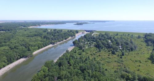 Aerial view of a river winding through lush greenery, leading to a large body of water under a clear blue sky.