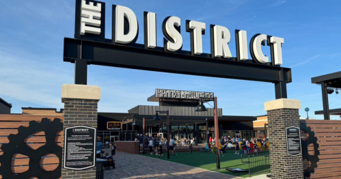 Entrance to The District, featuring a large sign and a bustling outdoor area with people enjoying the space.