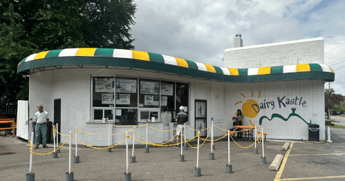 A retro-style ice cream shop with a colorful awning and outdoor seating, featuring a menu board and customers in line.