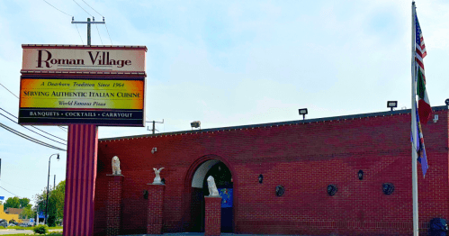 Sign for Roman Village restaurant, featuring Italian cuisine, with a brick building and American flag in the background.