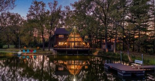 A cozy cabin with a triangular roof reflects in a calm pond at sunset, surrounded by trees and outdoor seating.