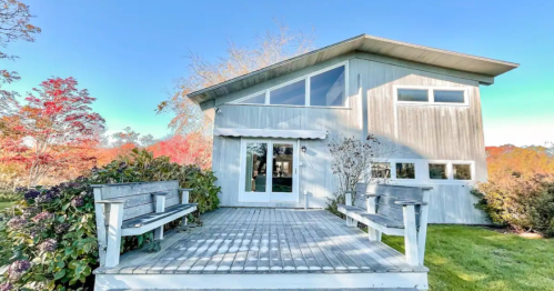 A modern two-story house with large windows, surrounded by colorful autumn foliage and a wooden deck with benches.