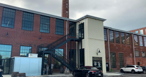 A brick building with large windows, a metal staircase, and a chimney, featuring a modern addition and parked cars.