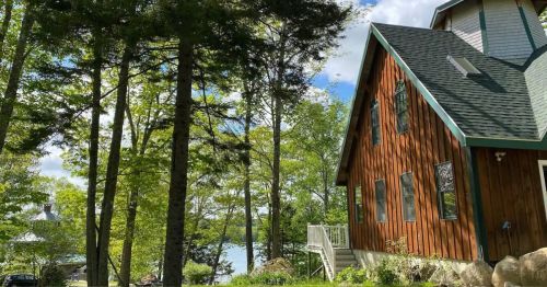 A rustic wooden cabin surrounded by trees, with a view of a lake in the background under a partly cloudy sky.