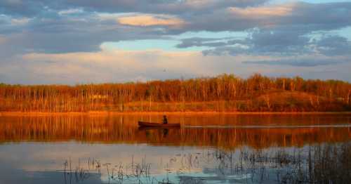 A lone person in a boat on a calm lake, surrounded by trees and reflecting colorful clouds at sunset.