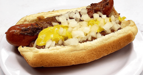 A hot dog in a bun topped with mustard, chopped onions, and chili on a white plate.