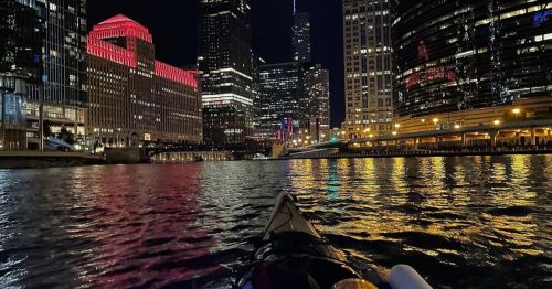 A kayak on a river at night, surrounded by illuminated skyscrapers reflecting on the water.