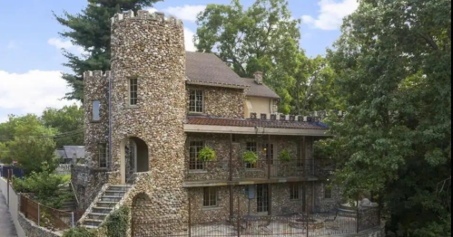A unique stone castle-style house with a turret, surrounded by trees and a fence.