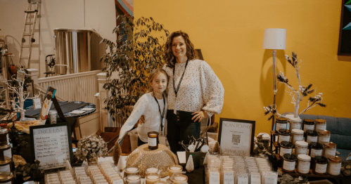 A woman and a girl stand behind a table filled with candles and products, smiling at the camera. Bright yellow wall in background.
