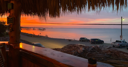 Sunset over a calm bay, with a pier in the distance and vehicles parked along the shore, viewed from a thatched-roof bar.