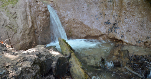 A small waterfall cascades over a rocky surface into a clear stream, surrounded by earthy tones and natural debris.