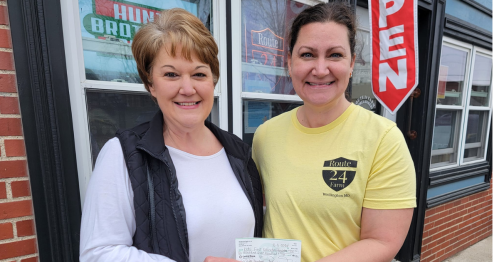Two women stand outside a shop, smiling and holding a check, with a red "OPEN" sign in the background.