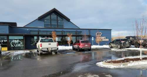 A modern blue building with large windows, featuring a sign for a restaurant, surrounded by parked cars and snow.