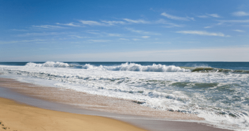 Waves crashing on a sandy beach under a clear blue sky with scattered clouds.
