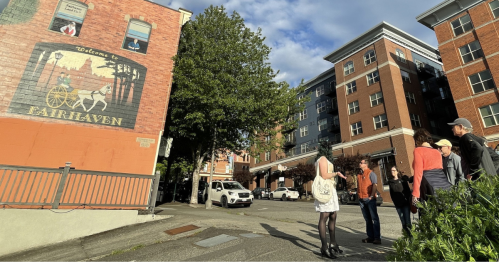A group of people stands on a sidewalk near a mural welcoming visitors to Fairhaven, with buildings and trees in the background.