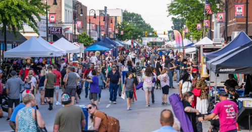 A bustling street fair with crowds, vendor tents, and colorful banners lining the road on a sunny day.