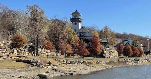 A scenic view of a lakeside with a lighthouse and autumn trees along the shore under a clear blue sky.