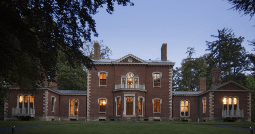 A historic brick mansion with large windows, surrounded by trees, illuminated at dusk.