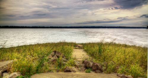 A pathway leads to a calm river, surrounded by tall grass and a cloudy sky in the background.