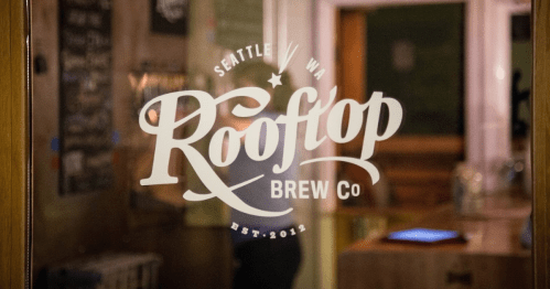 Sign for Rooftop Brew Co, located in Seattle, WA, established in 2012, seen through a glass door.