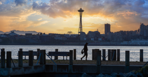 Silhouette of a person walking along a waterfront at sunset, with the Seattle skyline and Space Needle in the background.