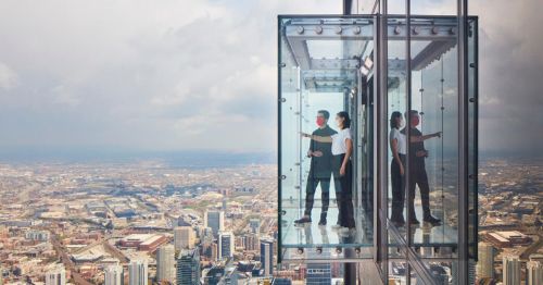 Two people in a glass elevator overlooking a city skyline from a tall building, with clouds in the background.