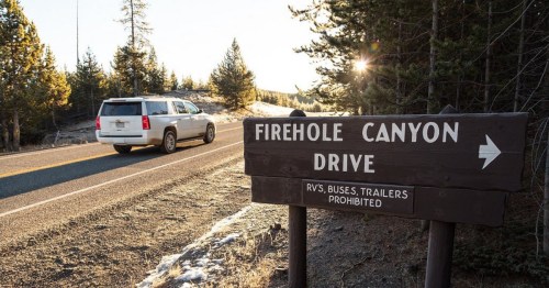 A vehicle drives past a wooden sign for Firehole Canyon Drive, surrounded by trees and sunlight.