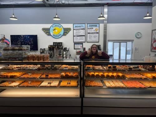 A smiling employee stands behind a display case filled with various donuts and pastries at a donut shop.