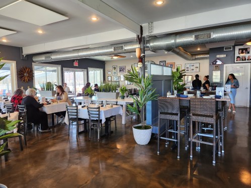 A bright restaurant interior with tables, plants, and patrons dining, featuring a bar area and wall decor.