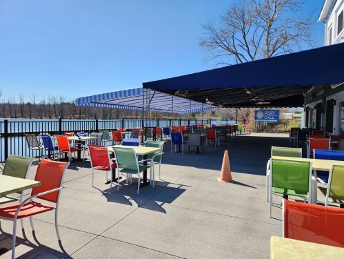 Outdoor dining area with colorful chairs and tables by a lake, under a blue sky and shaded by a large awning.
