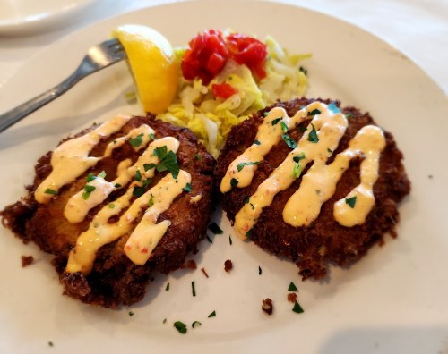 Two golden-brown crab cakes drizzled with sauce, served with a lemon wedge and a side of shredded lettuce and diced tomatoes.