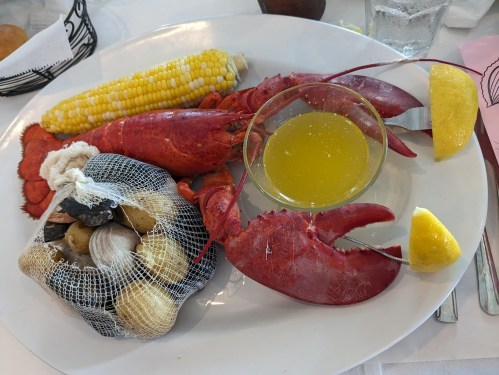 A plate featuring a lobster, corn on the cob, a seafood bag, and lemon wedges with a bowl of melted butter.