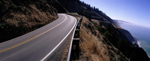 A winding road along a coastal cliff, with ocean views and lush greenery on the hillside.