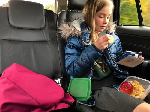 A girl in a blue jacket sits in a car, eating a snack from a container, with a backpack beside her.