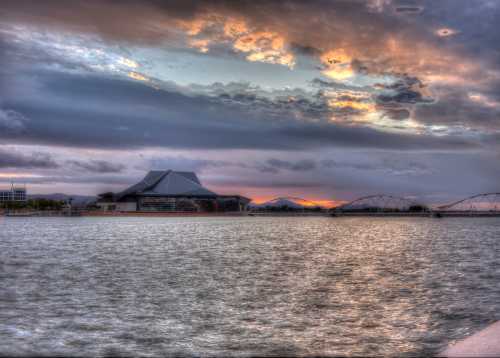 A scenic view of a river at sunset, featuring a modern building and a bridge against a colorful sky.