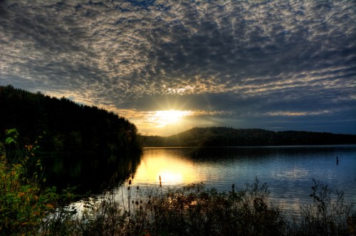 Sunset over a calm lake, with clouds reflecting in the water and trees lining the shore.