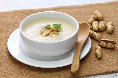 A bowl of creamy soup topped with peanuts, accompanied by a wooden spoon and scattered peanuts on a textured cloth.