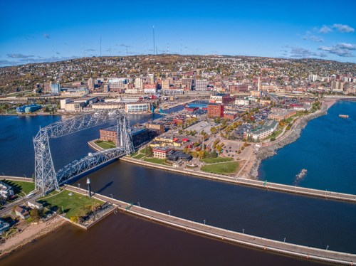 Aerial view of a city by the water, featuring a bridge, waterfront, and buildings against a clear blue sky.