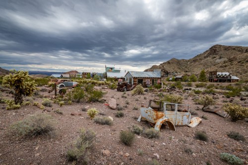 Abandoned ghost town with rusted cars, old buildings, and a dramatic cloudy sky in a desert landscape.